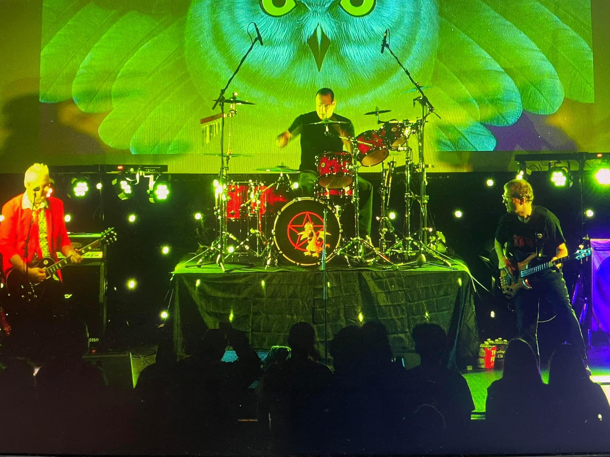 Driven, the band, in a gig, shot taken center stage of the whole band. The Rush Fly By Night album cover is on the video screen behind the band