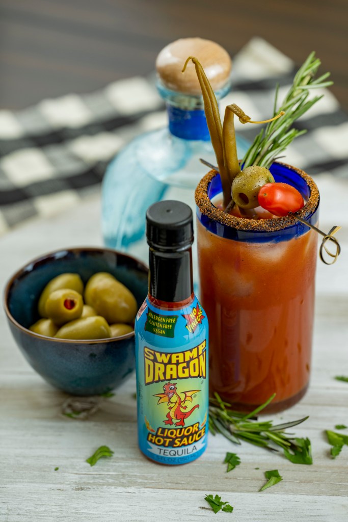 A bottle of Swamp Dragon's "Tequila Dragon" Hot Sauce next to a bloody mary coctail, garnished with pickled beans, an olive, and a cherry tomato